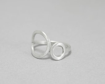 May include: A silver ring with two overlapping circles, one larger than the other. The ring has a brushed finish.
