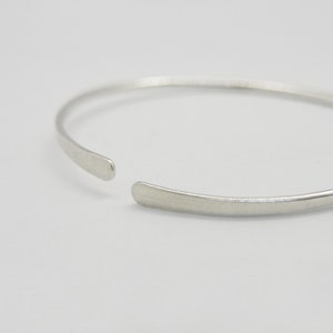 May include: A simple silver bangle bracelet with a slightly rounded shape. The bracelet is open on one side and has a smooth, polished finish.