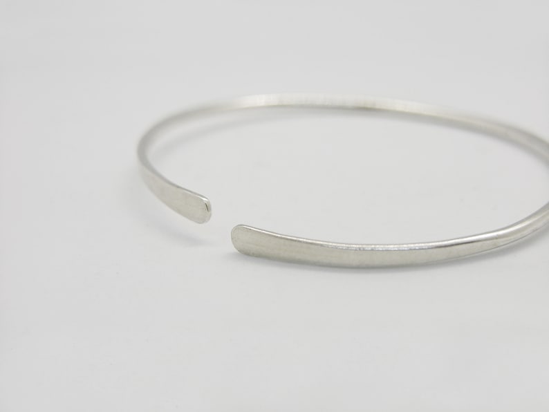 May include: A simple silver bangle bracelet with a slightly rounded shape. The bracelet is open on one side and has a smooth, polished finish.