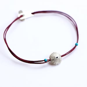 Könnte beinhalten: Ein Armband aus burgunderfarbenem Band mit einem silbernen Seeigel-Anhänger und türkisfarbenen Perlen.