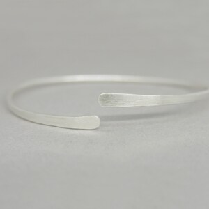 May include: A simple silver bangle bracelet with a slightly curved, open design. The bracelet is made of smooth, brushed metal.