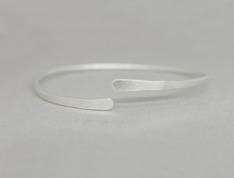 May include: A simple silver bangle bracelet with a slightly curved, open design. The bracelet is made of smooth, brushed metal.
