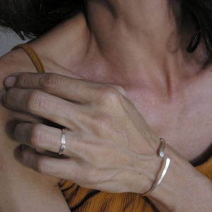 May include: A silver ring and a silver bangle bracelet on a person's hand. The ring is thin and has a simple design. The bangle is open and has a slightly curved shape.