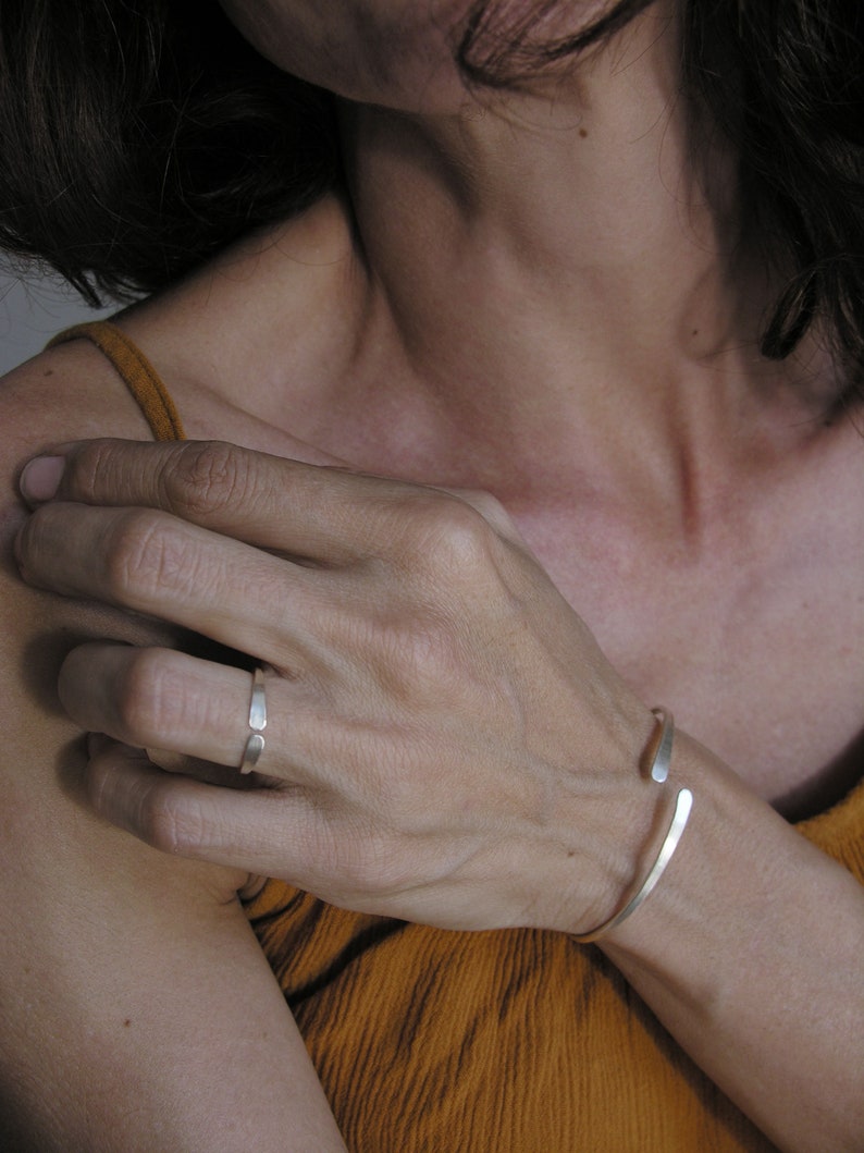 May include: A silver ring and a silver bangle bracelet on a person's hand. The ring is thin and has a simple design. The bangle is open and has a slightly curved shape.