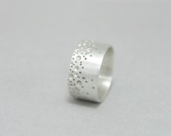 May include: A silver ring with a textured surface featuring a pattern of small, raised circles. The ring is wide and has a brushed finish.