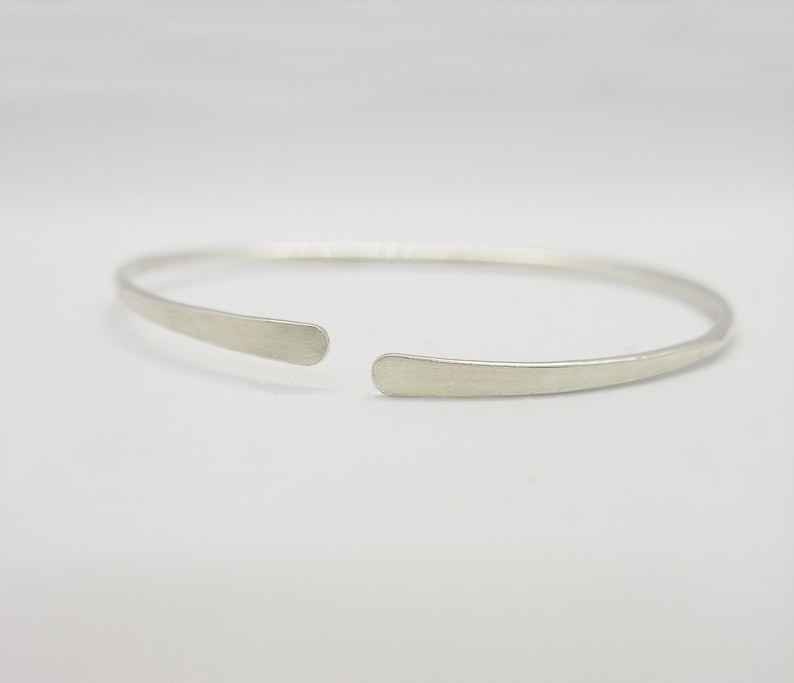 May include: A simple silver bangle bracelet with a hammered finish. The bracelet is open and has a slightly curved shape.