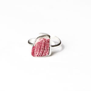 May include: A silver ring with a rectangular, pink stone with white veining. The stone is set in a silver bezel, and the ring band is a simple, open design. The ring is shown against a white backdrop.