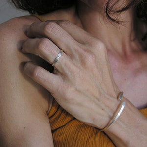 May include: A silver ring and bracelet on a person's hand. The ring is a simple band with a flat, rectangular shape. The bracelet is a thin, open band that wraps around the wrist.