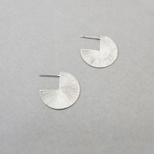 May include: A pair of silver earrings with a textured, geometric design. Each earring is a half-circle with a cut-out section.