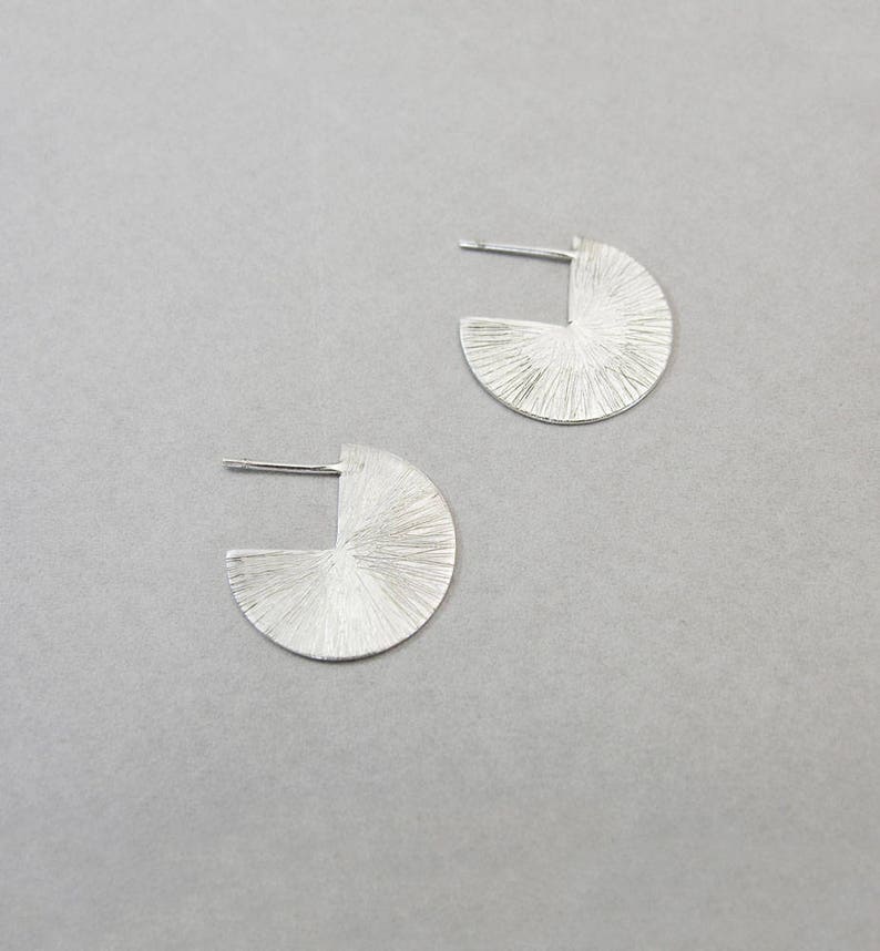 May include: A pair of silver earrings with a textured, geometric design. Each earring is a half-circle with a cut-out section.