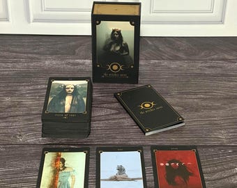 Puede incluir: Una baraja de cartas The Witches Tarot que incluye una caja negra con detalles dorados, un mazo de cartas y tres cartas individuales expuestas boca arriba. Las cartas muestran ilustraciones tituladas Seven of Cups, The Tower, Queen of Wands y The Moon.
