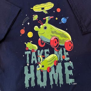 Puede incluir: Camiseta azul marino con un diseño caprichoso de vehículos con forma de pepinillo en el espacio. La frase "TAKE ME HOME" está impresa en una fuente goteante. El diseño incluye planetas y estrellas, creando una estética divertida y de dibujos animados.