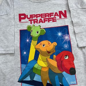 Puede incluir: Una camiseta gris claro de manga larga con un gráfico colorido de tres criaturas de dibujos animados y el texto "Pupperfan Traffe" en rojo. Las criaturas son verdes, naranjas y rojas con patas azules, sobre un fondo azul estrellado.