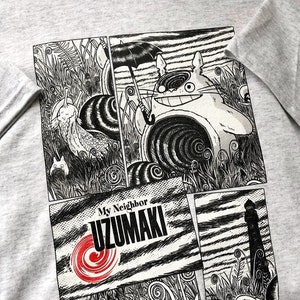 Puede incluir: Una camiseta gris con un gráfico en blanco y negro de estilo cómic de una criatura con una espiral en el ojo. El texto "My Neighbor Uzumaki" está impreso en rojo y blanco en la camiseta.