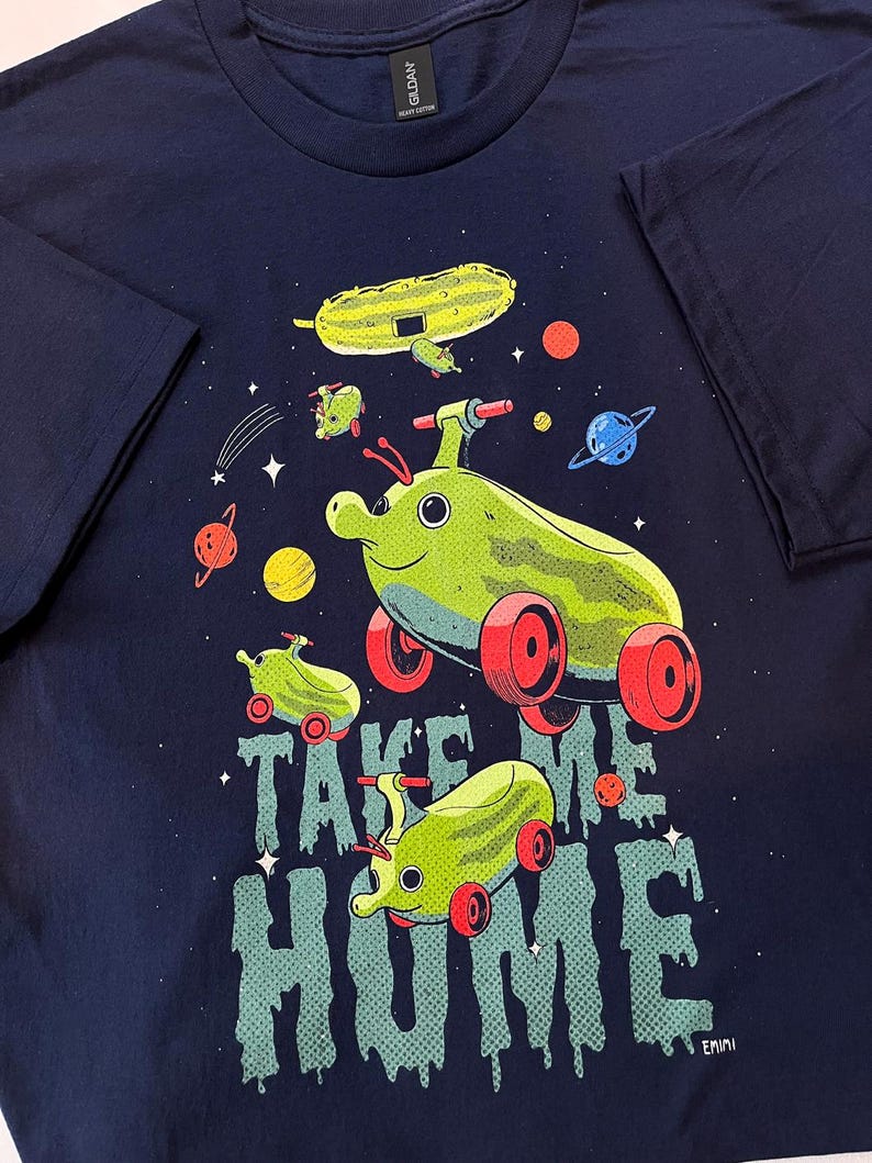 Peut inclure: T-shirt bleu marine avec un motif fantaisiste de v&eacute;hicules en forme de cornichons dans l'espace. L'expression "TAKE ME HOME" est imprim&eacute;e dans une police d&eacute;goulinante. Le motif comprend des plan&egrave;tes et des &eacute;toiles, cr&eacute;ant une esth&eacute;tique amusante et caricaturale.