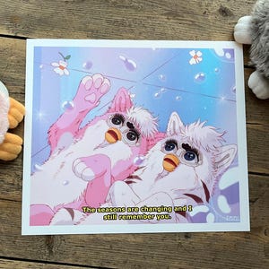 Puede incluir: Una impresión con dos criaturas estilizadas tipo Furby en un entorno azul y rosa con burbujas y flores. El texto "The seasons are changing and I still remember you" está en la parte inferior. Dos peluches están en el fondo.