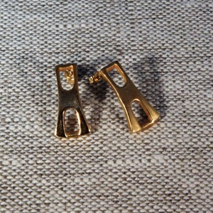 May include: A pair of gold-toned earrings with a geometric design. The earrings are shaped like a rectangle with a cutout in the middle.