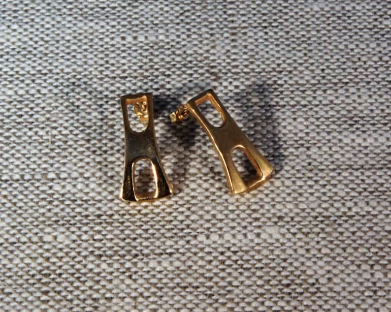 May include: A pair of gold-toned earrings with a geometric design. The earrings are shaped like a rectangle with a cutout in the middle.