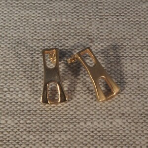May include: A pair of gold-toned earrings with a geometric design. The earrings are shaped like a rectangle with a cutout in the middle.
