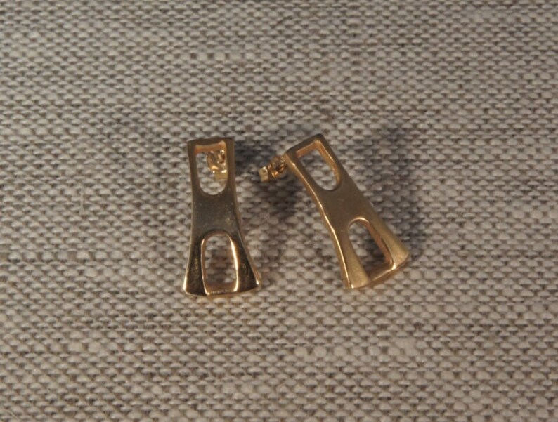 May include: A pair of gold-toned earrings with a geometric design. The earrings are shaped like a rectangle with a cutout in the middle.