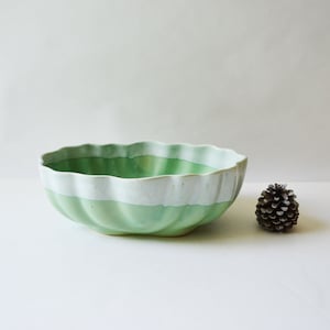 May include: A ceramic bowl with a scalloped rim, painted in shades of green and white. The bowl has a textured surface and a wide, shallow shape. A small pine cone sits to the right of the bowl. The bowl is likely used for serving food or as a decorative item.