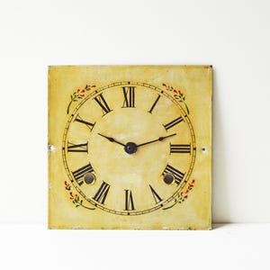 May include: Square clock face with black Roman numerals on a cream-colored background. The clock face has black hands and is decorated with small red and green floral accents. The clock face is square and has two small holes.