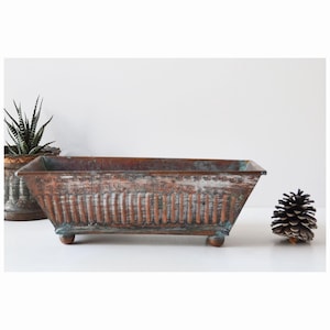 May include: A rectangular copper planter with a distressed finish and raised detailing. The planter has four small feet and is sitting on a white surface.