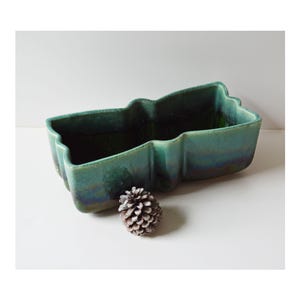 May include: A teal-colored ceramic planter with a unique, wavy design. The rectangular container has a glossy finish and is accompanied by a pinecone. The planter is suitable for indoor or outdoor use.