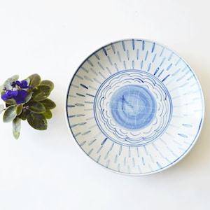 May include: A white ceramic plate with a blue and white design. The plate has a circular design in the center with a blue swirl and a border of blue lines. The plate is surrounded by a white background.
