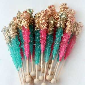 May include: A group of candy sticks with a teal, pink, and gold color scheme. The candy sticks are on wooden sticks with round wooden balls at the bottom.