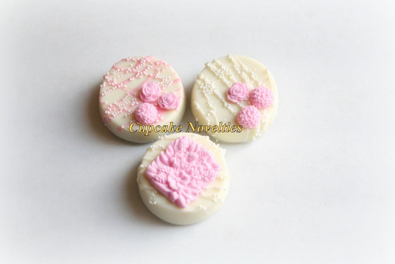 May include: Three white chocolate covered cookies decorated with pink frosting and white sprinkles. The cookies have different designs, including a floral pattern and a simple swirl.