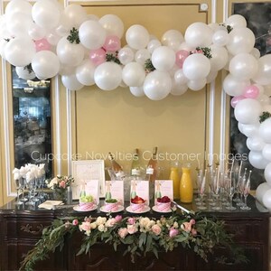 May include: A white and pink balloon arch with greenery decorates a bar with a floral garland, champagne bottles, and glasses. The bar is set up for a party or celebration.