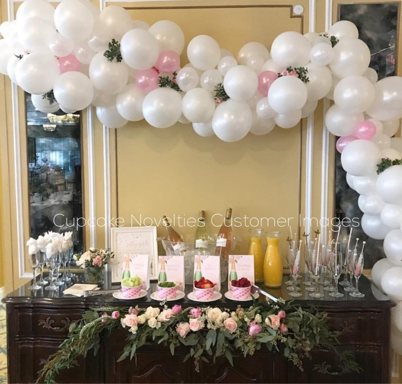 May include: A white and pink balloon arch with greenery decorates a bar with a floral garland, champagne bottles, and glasses. The bar is set up for a party or celebration.