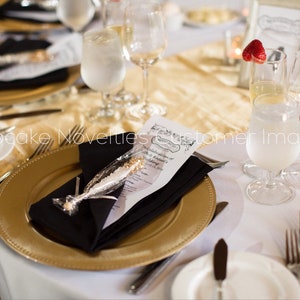 May include: A gold charger plate with a black napkin and a white favor bag with a gold and white candy stick. The plate is set on a white tablecloth with a gold runner. There are glasses and silverware on the table.