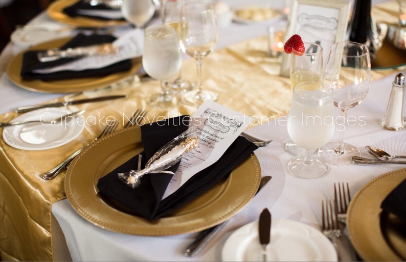 May include: A gold charger plate with a black napkin and a white favor bag with a gold and white candy stick. The plate is set on a white tablecloth with a gold runner. There are glasses and silverware on the table.