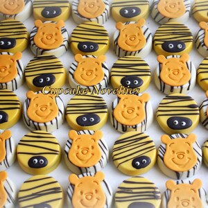 May include: A collection of round chocolate-covered cookies decorated with Winnie the Pooh's face and yellow and black stripes. The cookies are arranged in rows on a white surface. Cupcake Novelties is printed on the surface.