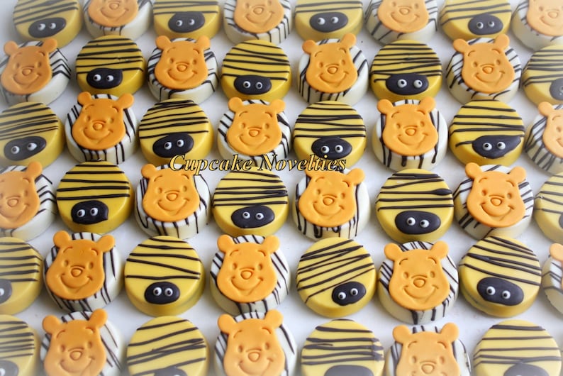 May include: A collection of round chocolate-covered cookies decorated with Winnie the Pooh's face and yellow and black stripes. The cookies are arranged in rows on a white surface. Cupcake Novelties is printed on the surface.