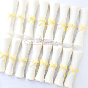 May include: A set of 18 white chocolate graduation diplomas with yellow ribbon bows. The diplomas are arranged in three rows of six. Cupcake Novelties is printed on the bottom of the image.