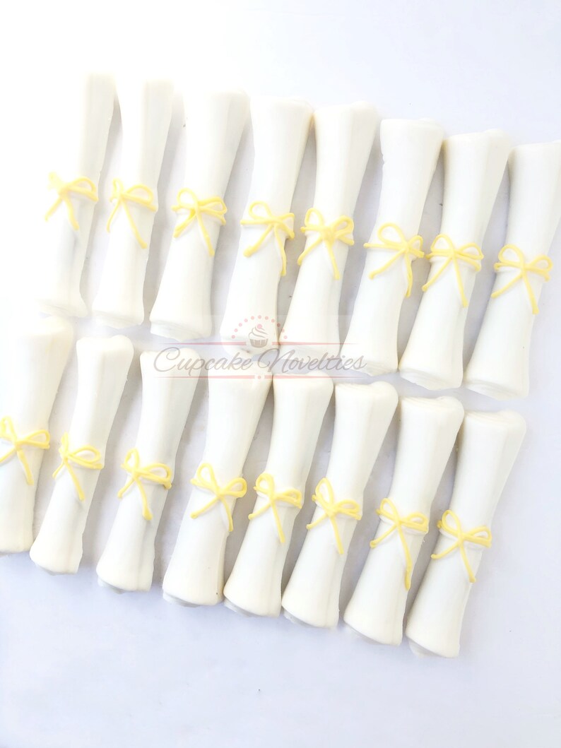 May include: A set of 18 white chocolate graduation diplomas with yellow ribbon bows. The diplomas are arranged in three rows of six. Cupcake Novelties is printed on the bottom of the image.