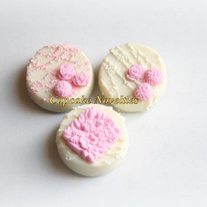 May include: Three white chocolate covered cookies decorated with pink frosting and white sprinkles. The cookies have different designs, including a floral pattern and a simple swirl.