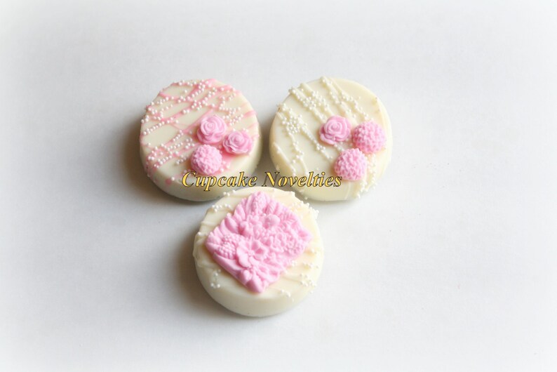 May include: Three white chocolate covered cookies decorated with pink frosting and white sprinkles. The cookies have different designs, including a floral pattern and a simple swirl.