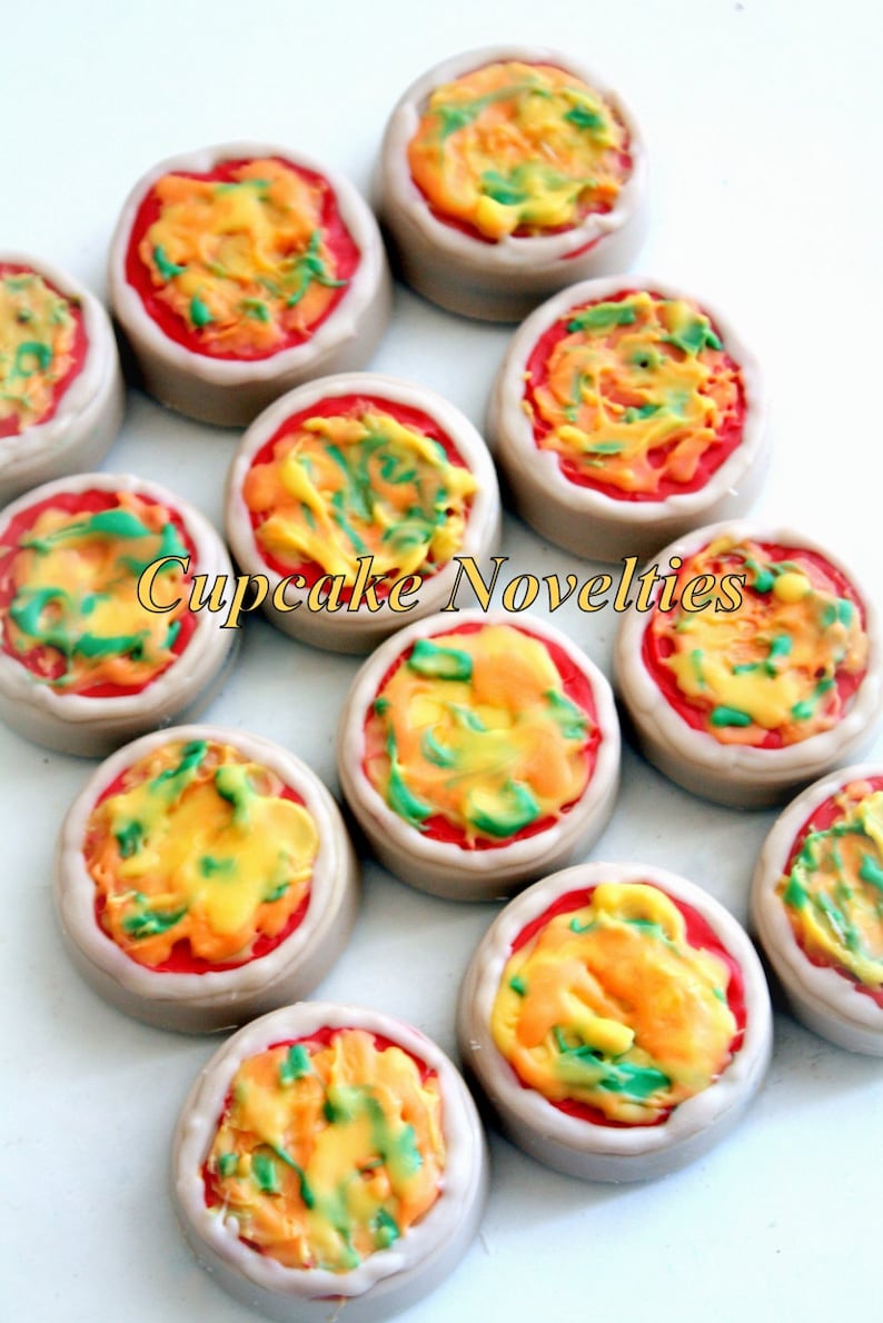 May include: Assortment of round, edible pizza-shaped cupcakes with red, yellow, and green frosting. The cupcakes are arranged in a circle on a white surface. The text "Cupcake Novelties" is visible in the center of the image.