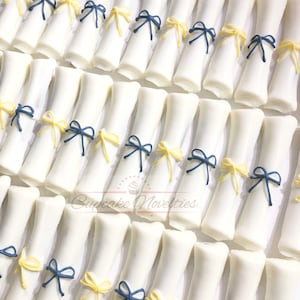 May include: White chocolate graduation cap shaped treats decorated with blue and yellow bows. Cupcake Novelties.