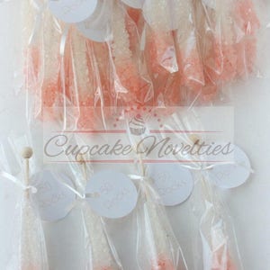 May include: A collection of small, clear plastic bags filled with pink and white rock candy. Each bag has a white circular tag attached with the text "50 Rocks". The bags are tied with white ribbon and a wooden stick. The image includes the text "Cupcake Novelties" and "Custom tags available for an extra fee".