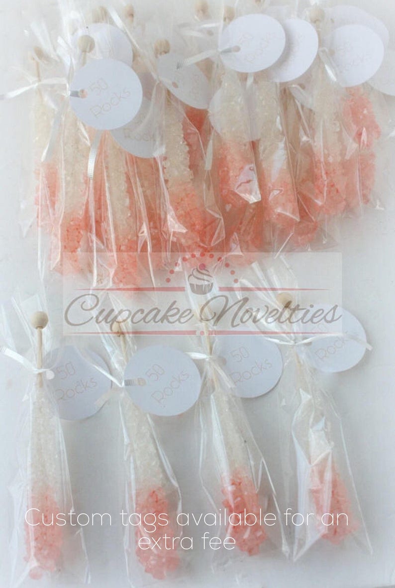 May include: A collection of small, clear plastic bags filled with pink and white rock candy. Each bag has a white circular tag attached with the text "50 Rocks". The bags are tied with white ribbon and a wooden stick. The image includes the text "Cupcake Novelties" and "Custom tags available for an extra fee".