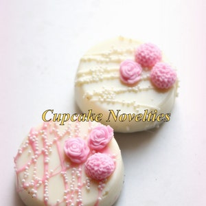 May include: Two white chocolate covered cookies decorated with pink flowers and white sprinkles.  The cookies are on a white background.  "Cupcake Novelties" is written in gold letters below the cookies.