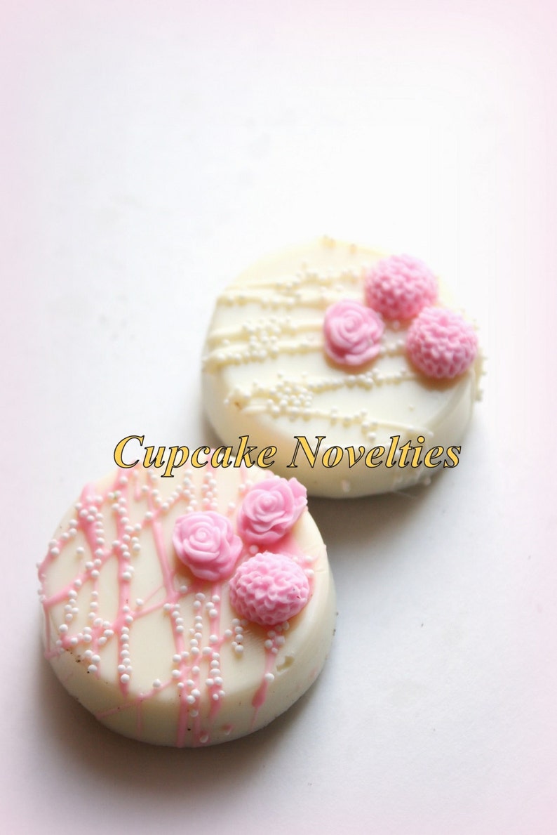 May include: Two white chocolate covered cookies decorated with pink flowers and white sprinkles.  The cookies are on a white background.  "Cupcake Novelties" is written in gold letters below the cookies.