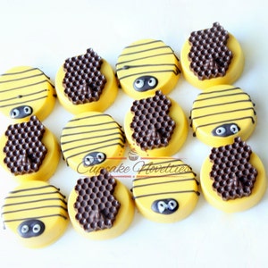 May include: A dozen yellow-frosted cookies decorated to resemble bees. Each cookie has black stripes, googly eyes, and a chocolate honeycomb design. The cookies are arranged on a white surface.