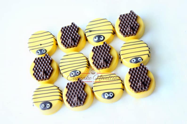 May include: A dozen yellow-frosted cookies decorated to resemble bees. Each cookie has black stripes, googly eyes, and a chocolate honeycomb design. The cookies are arranged on a white surface.