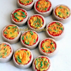 May include: A collection of 10 mini pizza cupcakes decorated with red, yellow, and green frosting. The cupcakes are arranged in a circle on a white surface. Cupcake Novelties is written in gold letters on the image.